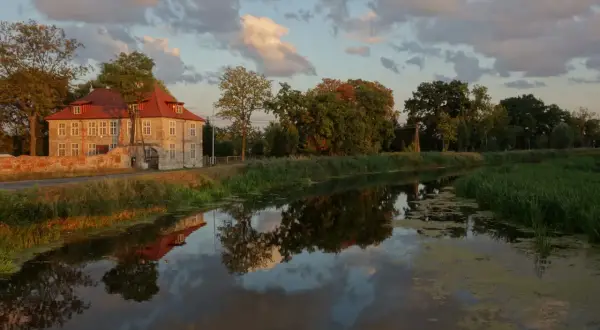 Dworek w Gdańsku Olszynce z czerwonym dachem odbija się w spokojnej wodzie kanału. Chmury na niebie malują zachód słońca.
