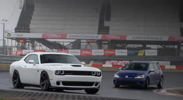 Biały Dodge Challenger i niebieski Volkswagen Golf na torze Słomczyn. W tle trybuny i banery reklamowe.