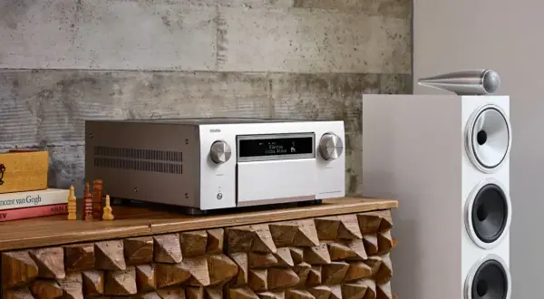 Srebrny amplituner Denon z wyświetlaczem Dolby Atmos, obok białej kolumny głośnikowej. To centrum domowego kina, które pozwala cieszyć się kinową jakością dźwięku.