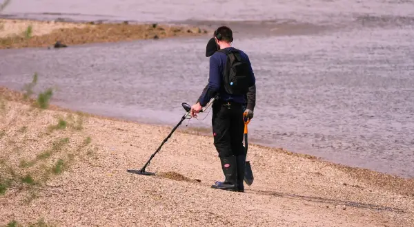 Mężczyzna z wykrywaczem metali i łopatką na plaży przy wodzie.