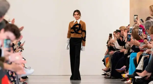 Victoria Beckham går på catwalken, klädd i en brun tröja med ett svart mönster och vida svarta byxor. Publiken, inklusive Harper Beckham, fotograferar med sina mobiler.
