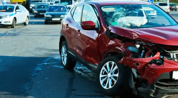 Wypadek drogowy. Czerwony samochód po kolizji, zniszczony przód i pęknięta szyba. Na drodze inne pojazdy, w tym radiowóz. Może to być sytuacja wymagająca **umowy na zastępstwo** kierowcy.