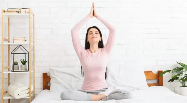 Mujer meditando en la cama, con las manos juntas sobre la cabeza, practicando dinámicas de relajación.