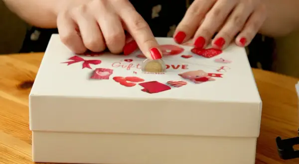Dłonie z czerwonymi paznokciami wkładają monetę do pudełka z napisem "Gift LOVE".