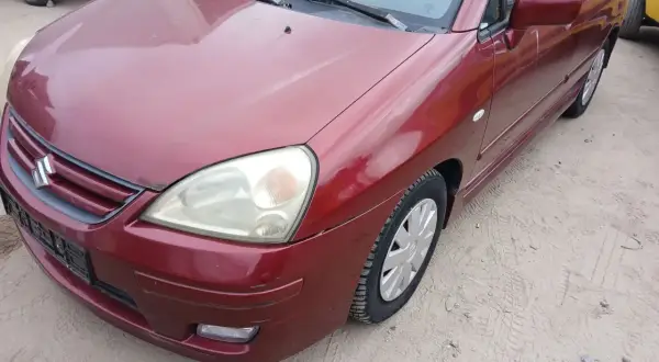 Czerwony Suzuki Liana stoi na piaszczystym podłożu. Widoczny przód auta z reflektorem i grillem.