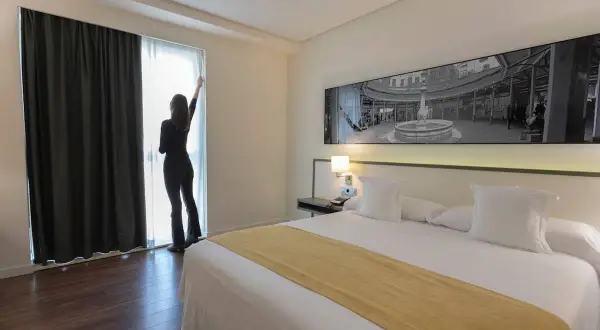 Habitación moderna en el spa hotel Primus Valencia. Una mujer abre las cortinas, revelando una cama impecable y una vista urbana.