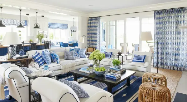 A bright living room with coastal interior design, featuring white sofas, blue and white patterned pillows, and a dark wood coffee table.