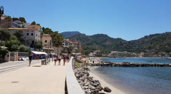 Promenad längs stranden i Port de Sóller med folk som njuter av solen och havet, omgivet av gröna berg.