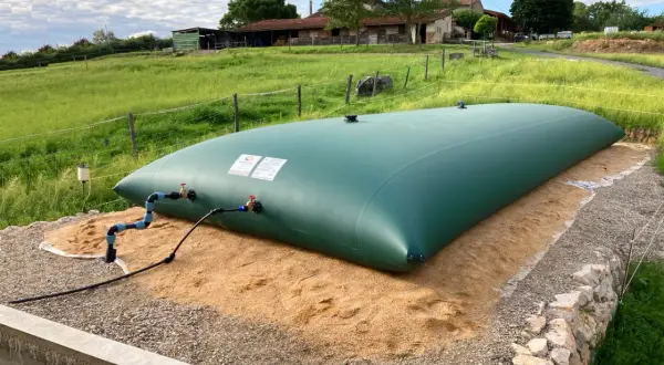 Gran depósito de agua de 20 litros, verde, instalado en un campo con una granja al fondo.