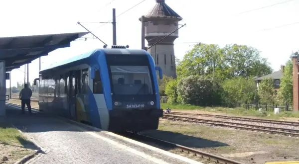 Niebieski pociąg SA103-010 czeka na pasażerów na peronie. W tle stara wieża ciśnień i drzewa. Wygodny wicie dojazd PKP.