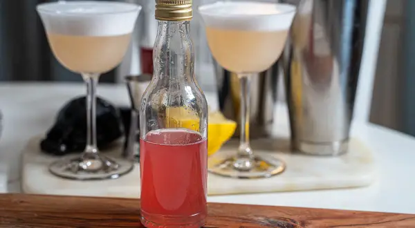 En flaska med rosa rabarbersirap står framför två drinkar med skum. En shaker och en citronklyfta syns i bakgrunden.