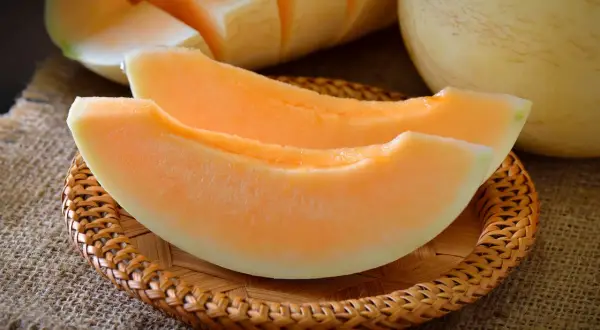 Dwa soczyste kawałki melona, jakby w ciąży, leżą na plecionej podstawce. W tle widać resztę owocu.