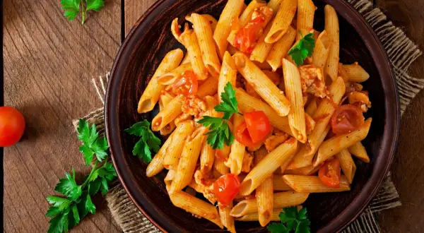 Talerz penne z pomidorami i kurczakiem, udekorowany natką pietruszki, na drewnianym tle.