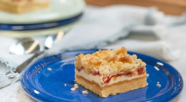 En bit rabarber cheesecake med smuldeg på en blå tallrik.