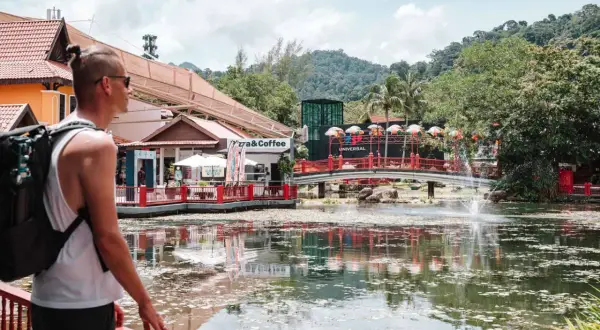 Mann mit Rucksack, der die bunte Malaysia Kleidung und die tropische Landschaft genießt. Ein See mit Fontäne und Brücke.