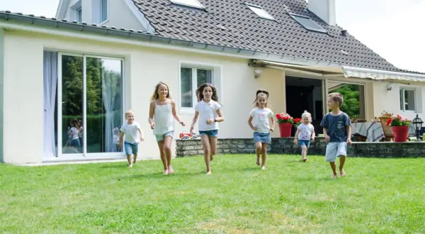 Dzieci biegają po trawniku przed domem z jasnym tynkiem zewnętrznym. Różne rodzaje tynków zewnętrznych mogą nadać budynkowi unikalny wygląd.