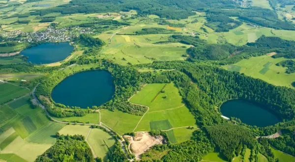 Luftaufnahme des **rundwegs um die drei Dauner Maare**. Grüne Felder, Wälder und drei dunkle Maare prägen die Landschaft.