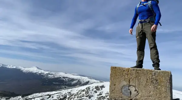 Hombre con mochila en la cima de un pico nevero, con vistas a montañas nevadas bajo un cielo azul con nubes.