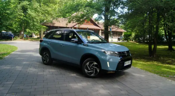 Jasnoniebieski Suzuki Vitara Hybrid zaparkowany na wybrukowanym podjeździe, otoczony zielenią i drewnianym budynkiem.