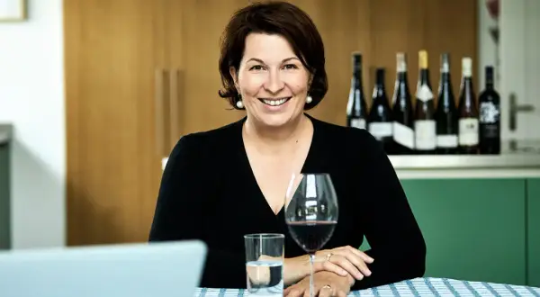 Eine lächelnde Frau, die wie ein **master of wine** wirkt, sitzt an einem Tisch mit Wein und Wasser. Im Hintergrund stehen mehrere Weinflaschen.