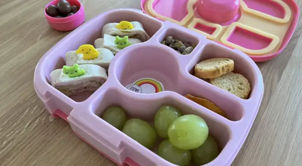 Różowa śniadaniówka do szkoły z przegródkami zawiera zawinięte tortille z uroczymi ozdobami, winogrona, orzeszki pistacjowe, krakersy i suszone mango.