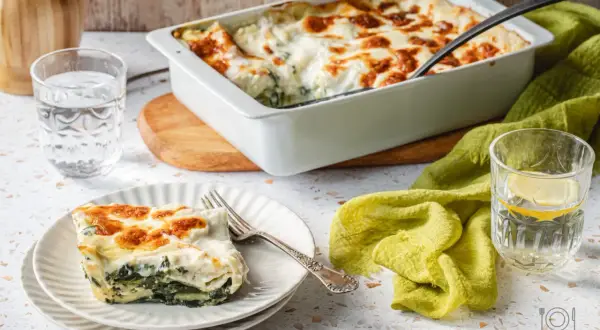 Pyszne lasagne ze szpinakiem, zapieczone na złoto, podane na talerzu obok szklanki wody z cytryną.