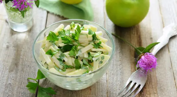 Frischer Kohlrabi Salat mit Petersilie in einer Glasschüssel, garniert mit einer lila Blume und grünen Äpfeln im Hintergrund.
