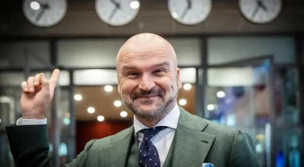 Rafał Brzoska, z uśmiechem i uniesionym palcem, symbolizuje swój rosnący majątek. Za nim zegary pokazują różne strefy czasowe.