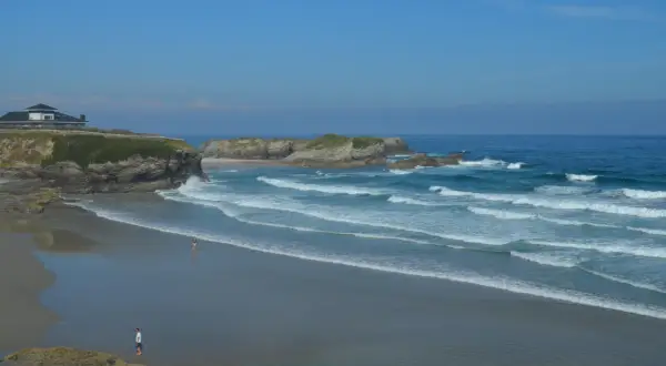 La **playa Os Castros** se extiende bajo un cielo azul, con olas rompiendo en la arena dorada. Una casa moderna corona el acantilado.