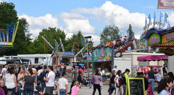 Buntes Treiben auf einem Volksfest mit Fahrgeschäften und vielen Besuchern. Ein Schiff-Karussell dreht sich, während Menschen an Ständen vorbeischlendern.