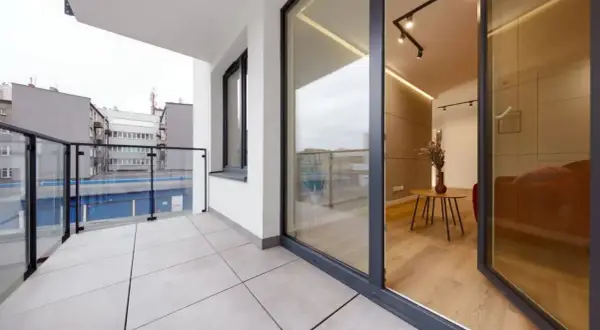 Nowoczesny balkon z płytkami, szklaną balustradą i widokiem na budynki.