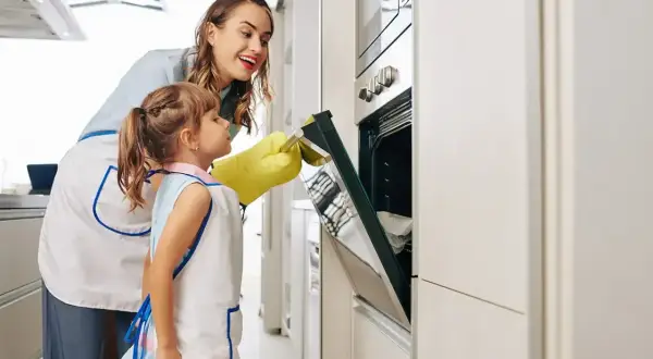 Mama i córka w fartuszkach otwierają piekarnik, by sprawdzić, czy trwa rozmrażanie w piekarniku.