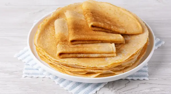 Pile de crêpes dorées, certaines pliées, sur une assiette blanche, posée sur une serviette à rayures bleues. Une délicieuse crêpe sans oeuf prête à être dégustée.