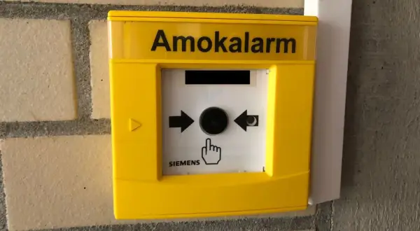Gelber Amokalarm-Knopf an einer Ziegelwand. Ein Symbol zeigt eine Hand, die auf den Knopf drückt, um einen Amoklauf an einer Schule zu melden.