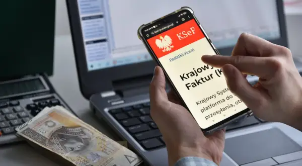 Dłoń trzyma smartfon z ekranem KSeF, obok leżą pieniądze i kalkulator.