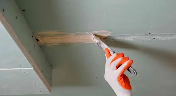 Ręka w rękawicy nakłada masę szpachlową na połączenie płyt gk, przygotowując je do klejenia na grzebień.