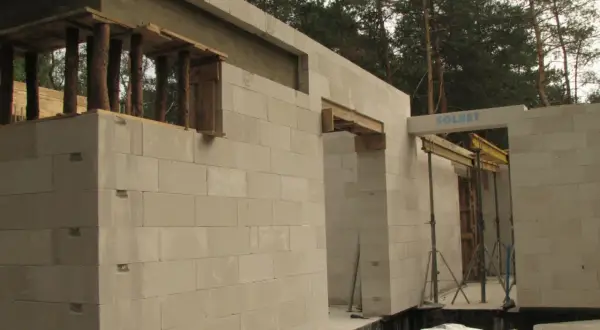 Budowa domu z bloczków betonowych. Widoczna ściana nośna z otworami okiennymi i drzwiami, podparta drewnianymi belkami.