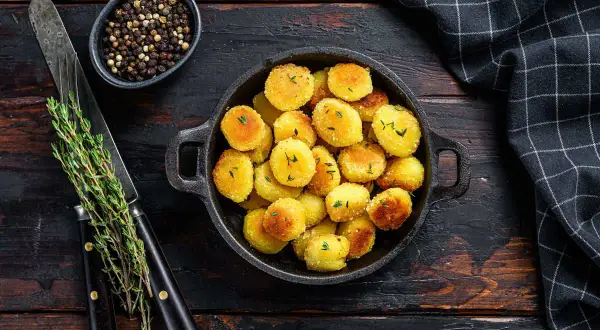 Crispy golden gnocchi in a black pan, garnished with herbs, on a wooden table with peppercorns and a knife.