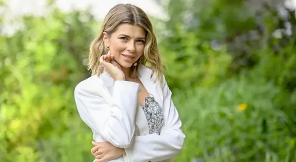 Agnieszka Dziekan z Koła Fortuny, uśmiechnięta, w białej marynarce, na tle zieleni.