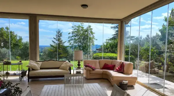 Soggiorno luminoso con divani in pelle e tessuto, tavolo in legno e ampie vetrate che incorniciano un panorama verde. Idee case con vetrate per godersi la natura.