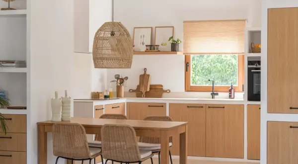 Cocina estilo mediterráneo con mesa de madera, sillas de mimbre y armarios de roble. Lámpara de mimbre y ventana con vistas a la vegetación.