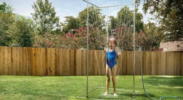 Ein Mädchen im blauen Badeanzug genießt eine erfrischende Dusche unter einer selbstgebauten Gartendusche im sonnigen Garten.