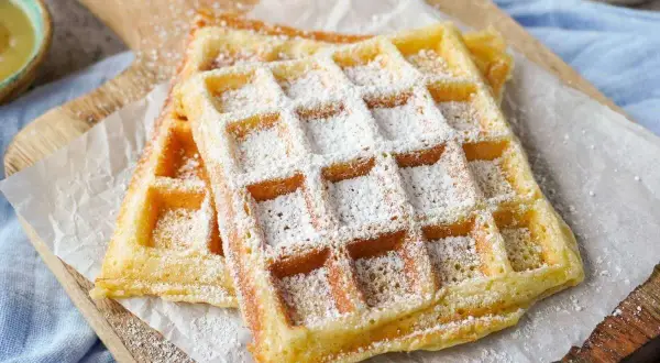 Zwei fluffige Waffeln, bestäubt mit Puderzucker, auf einem Holzbrett. Ein perfektes Rezept für fluffige Waffeln!