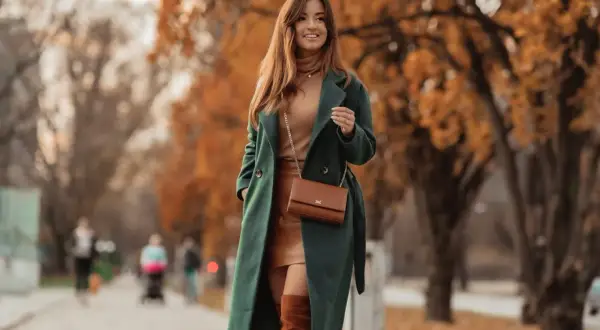 Elegancki, zielony płaszcz z bukli otula kobietę podczas jesiennego spaceru. W tle złote liście drzew.