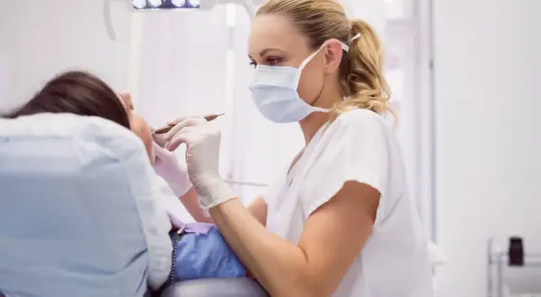 Dentystka w masce i rękawiczkach bada pacjentkę, która leży na fotelu. Może to być kontrola po zabiegu, np. po zerwanych szwach na dziąśle.