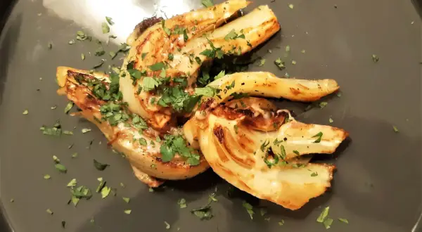 Złocisty, podsmażony koper włoski, posypany świeżą natką pietruszki, prezentuje się apetycznie na szarym talerzu.