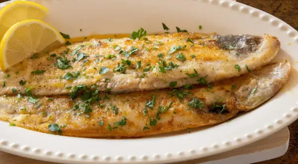 Delicioso lenguado a la plancha, dorado y jugoso, adornado con perejil fresco y rodajas de limón.