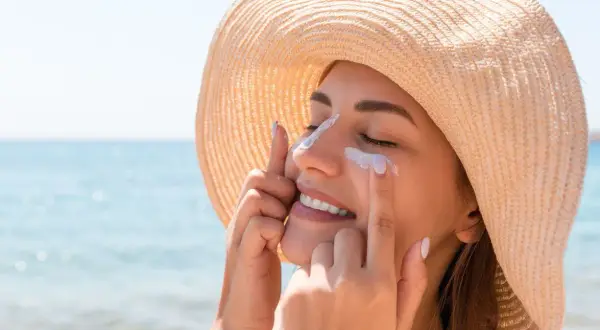 Une femme souriante applique de la crème solaire sur son visage, protégeant sa peau. Est-ce que la crème solaire hydrate ? Oui, certaines le font !