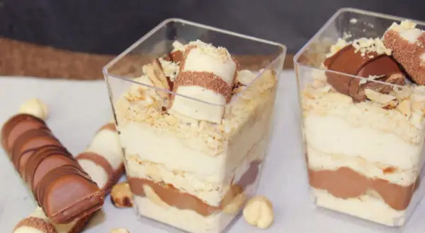 Deser Kinder Bueno w pucharkach z warstwami kremu, ciastek i czekolady, ozdobiony kawałkami batonika i orzechami.