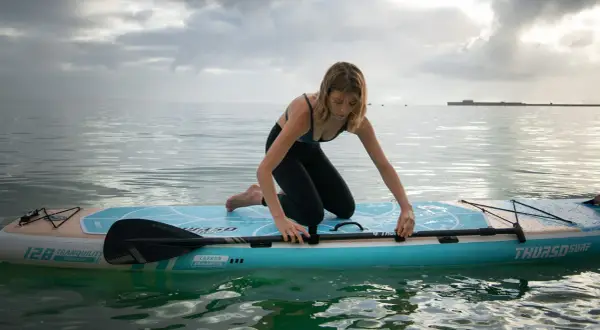 Donna in ginocchio su un sup, pronta a pagaiare. La tavola galleggia su acqua calma sotto un cielo nuvoloso.